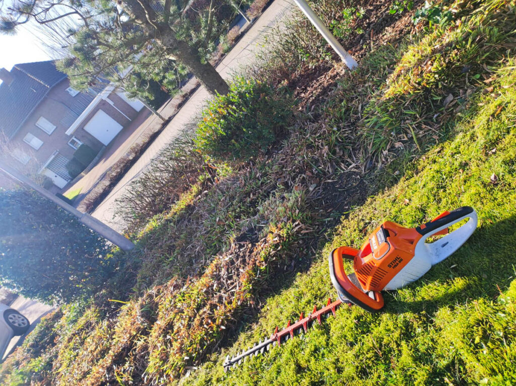 Eine motorisierte Heckenschere der Marke Stihl liegt auf dem Rasen vor einem frisch geschnittenen Beet mit Bodendeckern und anderen Pflanzen.